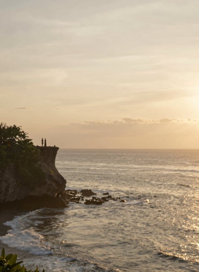 A wide-angle, serene shot of the Bali coastline at golden hour. The sky is a soft blend of #F7F3EE and muted gold. In the distance, the silhouette of a couple stands on a cliff edge, capturing a sense of scale and peace. The lighting is warm and ethereal, reflecting the brand's heartfelt elegance.