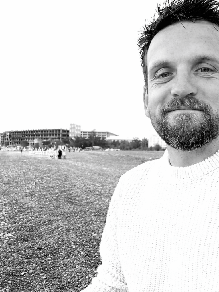 A bearded man in a white sweater smiling on a rocky beach in a black and white portrait.