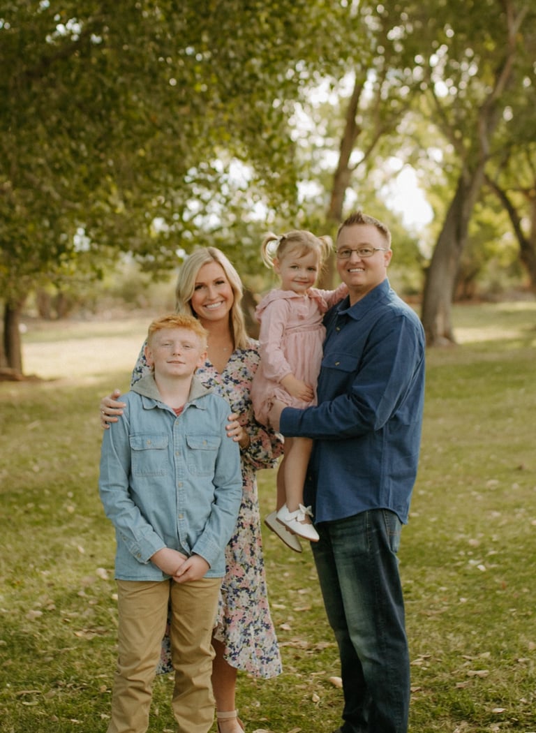 A happy family posing for an outdoor portrait in a grassy park surrounded by green trees.