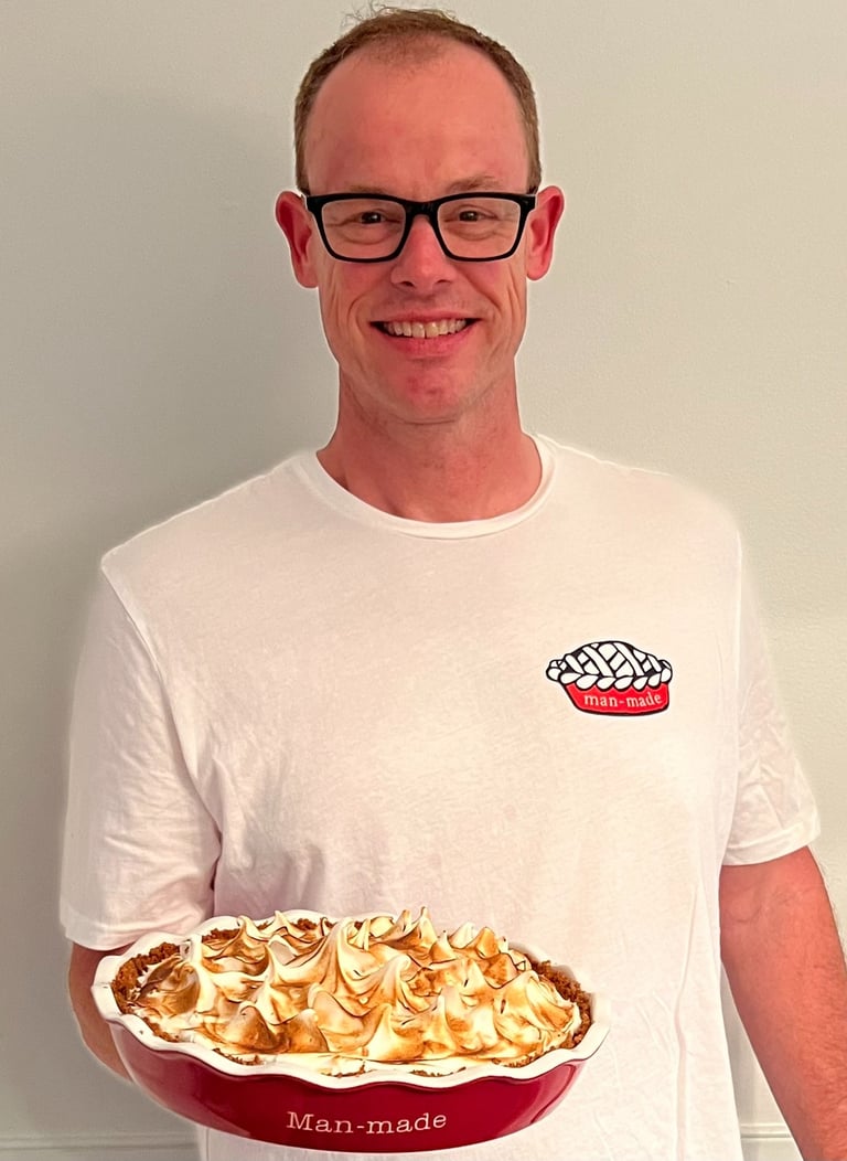 A smiling man wearing glasses holds a freshly baked lemon meringue pie in a red Man-made dish.