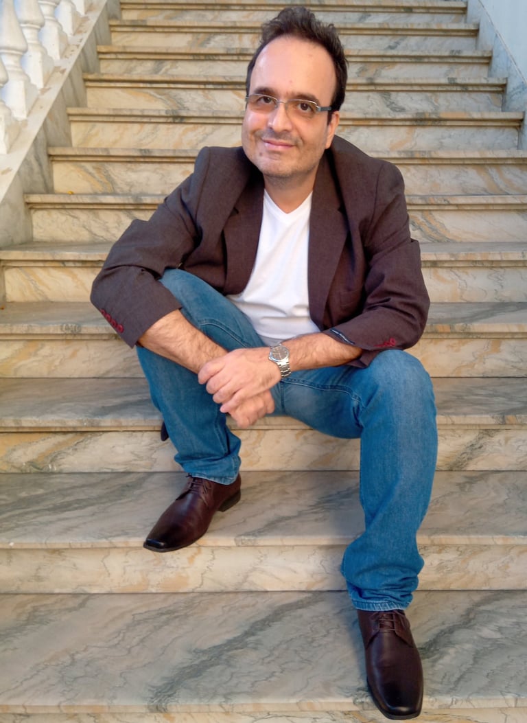 Jornalista Marcelo Dias, sentado à uma escadaria de mármore, de calça jeans, camisa branca e blazer.