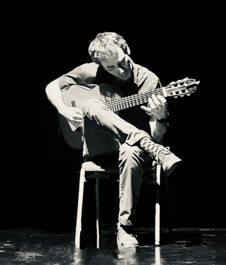 Paco playing the guitar under a theatrical overhead light.