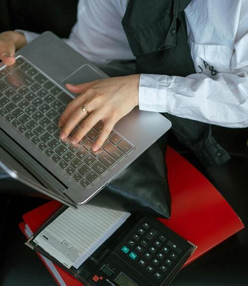 Una persona escribe en una laptop plateada junto a una calculadora y una carpeta roja
