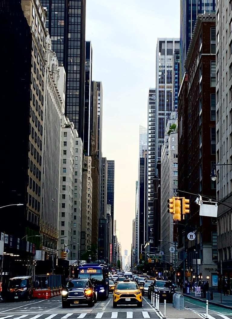 A bustling New York City streetscape with tall skyscrapers, traffic, and pedestrians under a bright sky.