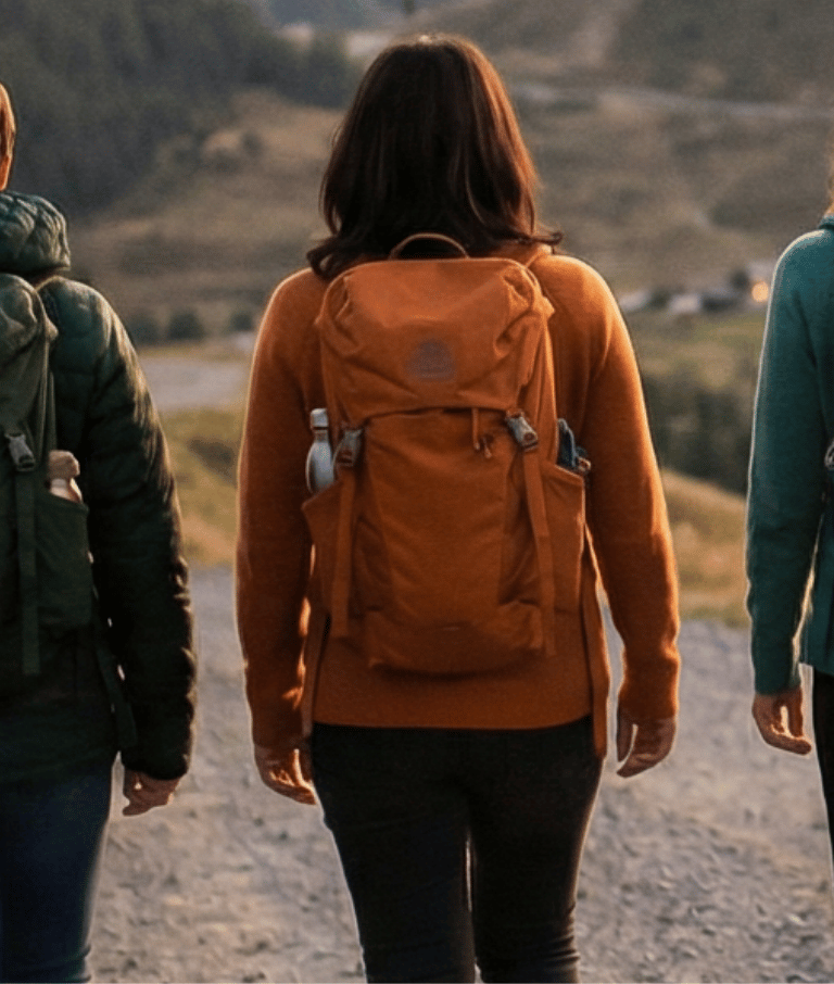 Four women with backpacks walk along a gravel road through rolling hills at dusk, seen from behind.