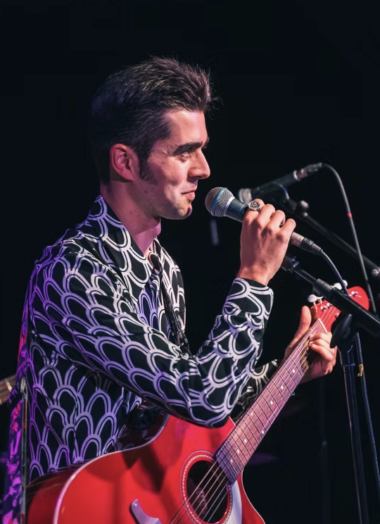 Olly Williams on stage in Brighton, focused while playing guitar and performing into a microphone.
