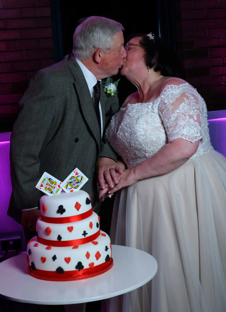 A bride and groom kiss behind a deck of cards themed wedding cake at their reception.