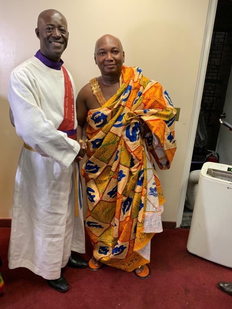 A smiling priest in a white alb standing next to a man wearing a traditional colorful Ghanaian Kente cloth.