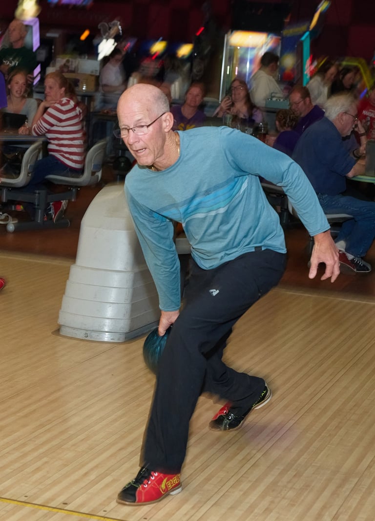 A senior man bowling in a teal shirt and black pants at a bowling alley with people in the background.
