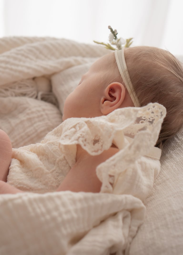 Newborn baby girl sleeping peacefully in a lace outfit and floral headband on a soft white blanket.