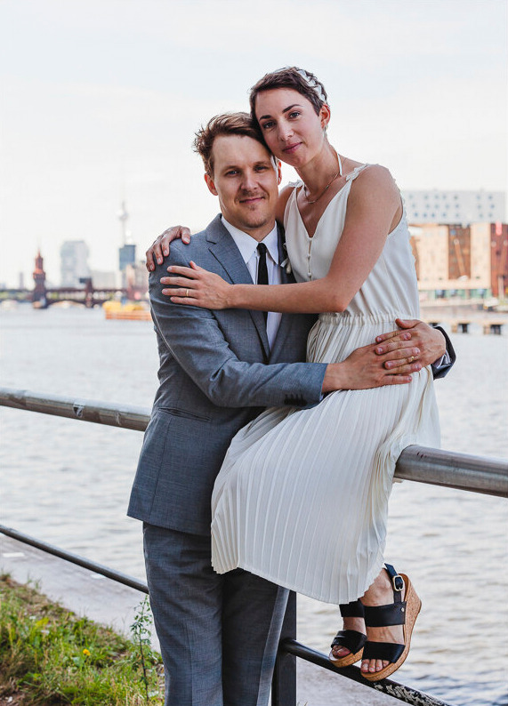 Bride sitting on a railing next to a river in Berlin holding her husband.