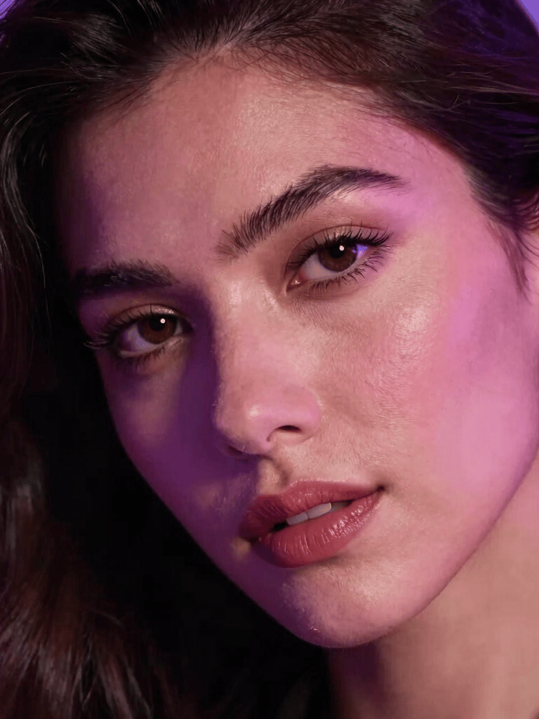 Close-up photograph of an adult model looking confidently into the camera, glowing under soft purple and pink neon highlights, deep shadows, luxurious global aesthetic, Latin American / Global style.