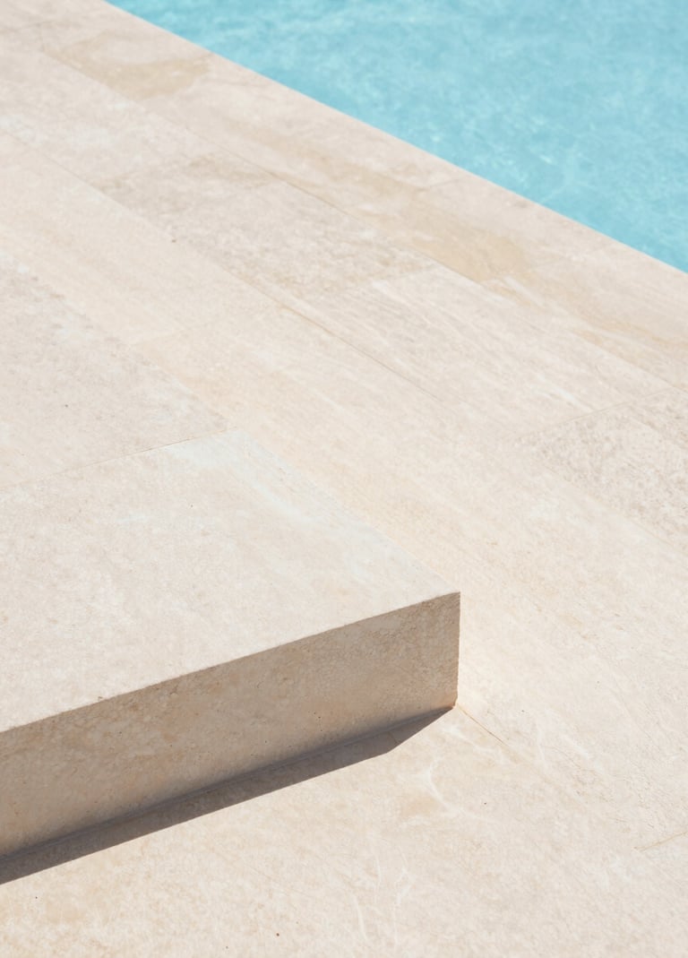 A close-up architectural detail of a minimalist stone terrace in Portugal. Off-white stone slabs meet the edge of a light blue infinity pool. Precise lines, clean composition, bright daylight.