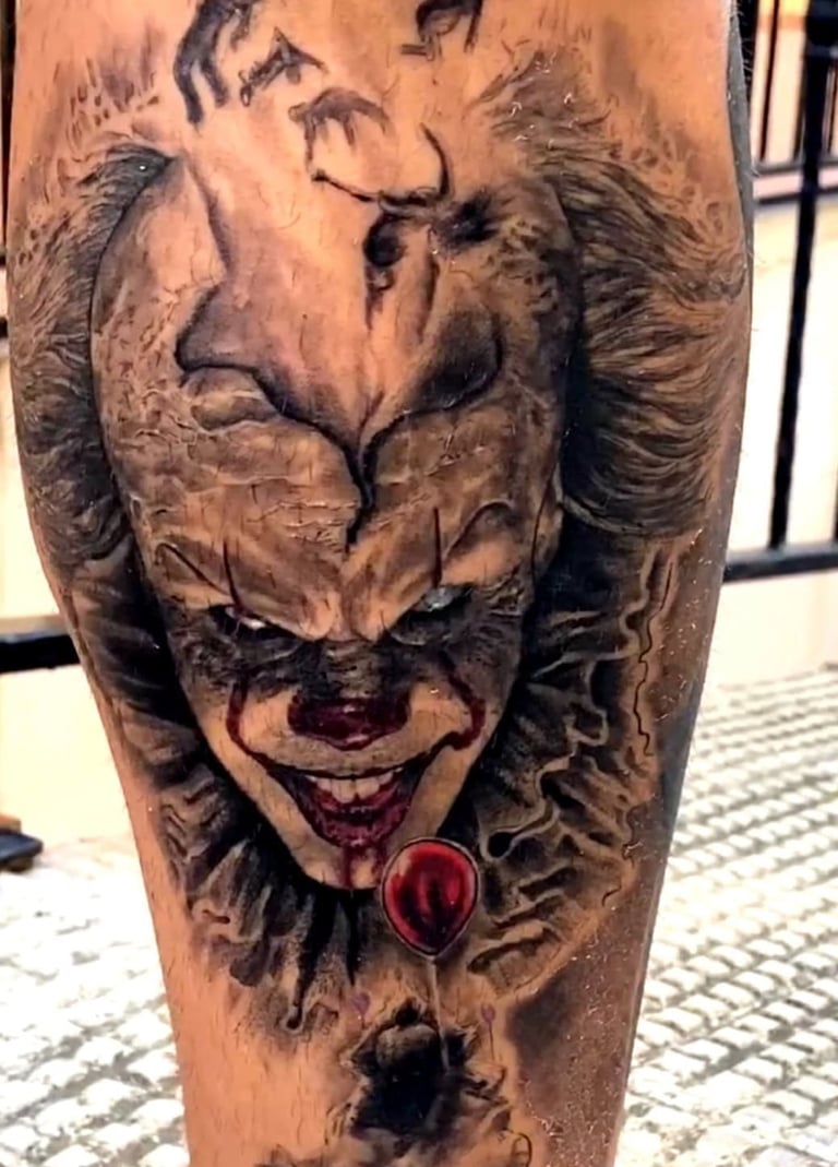 A realistic black and grey Pennywise the clown tattoo with a red balloon on a person's leg.