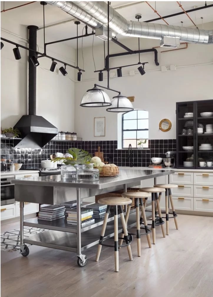 A large stainless steel table on wheels used as a kitchen island with bar stools and a lower shelf for cookbooks.