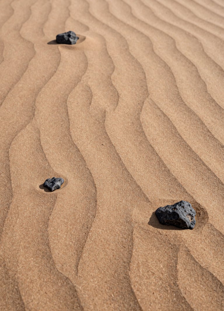 Detailed photographic shot of natural sand ripples and small desert rocks. The lighting is soft and warm, highlighting the natural beauty of the textures. Colors include soft sand, tan, and charcoal.
