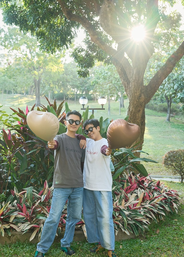 A happy couple wearing sunglasses holds heart-shaped balloons in a sunny park at sunset.
