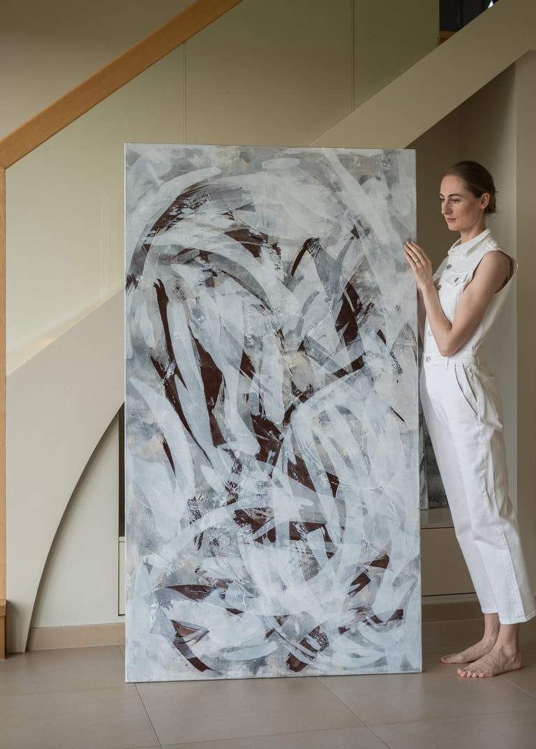 A female artist in white attire presents a large abstract painting in a modern home interior.