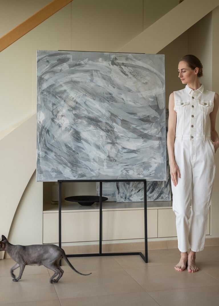 A woman in a white jumpsuit stands by abstract canvas art and a Sphynx cat in a modern home.