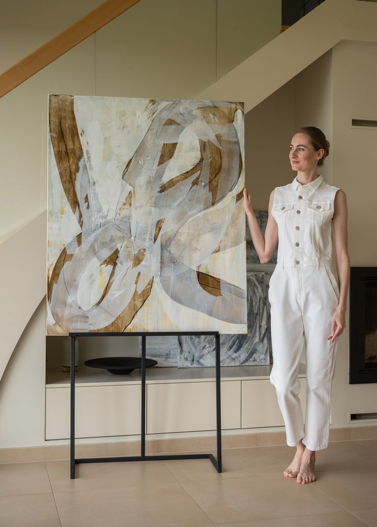 Artist in a white jumpsuit posing with a large abstract canvas painting in a modern home.
