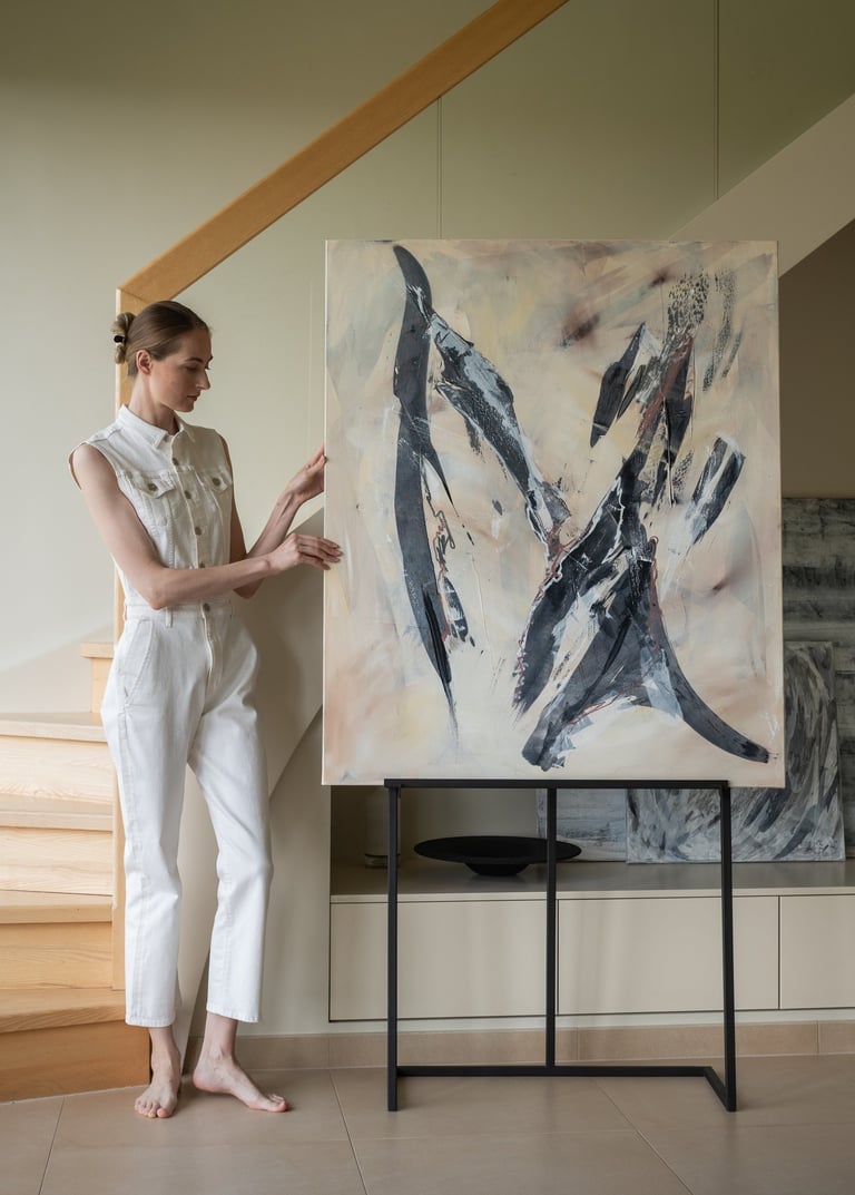 A female artist in a white jumpsuit poses beside a large abstract canvas painting on an easel in a modern minimalist home.