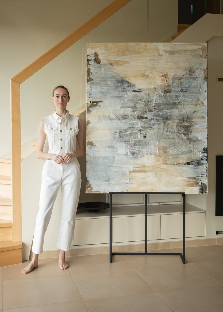 A female artist in a white jumpsuit stands by a large abstract painting in a modern home.