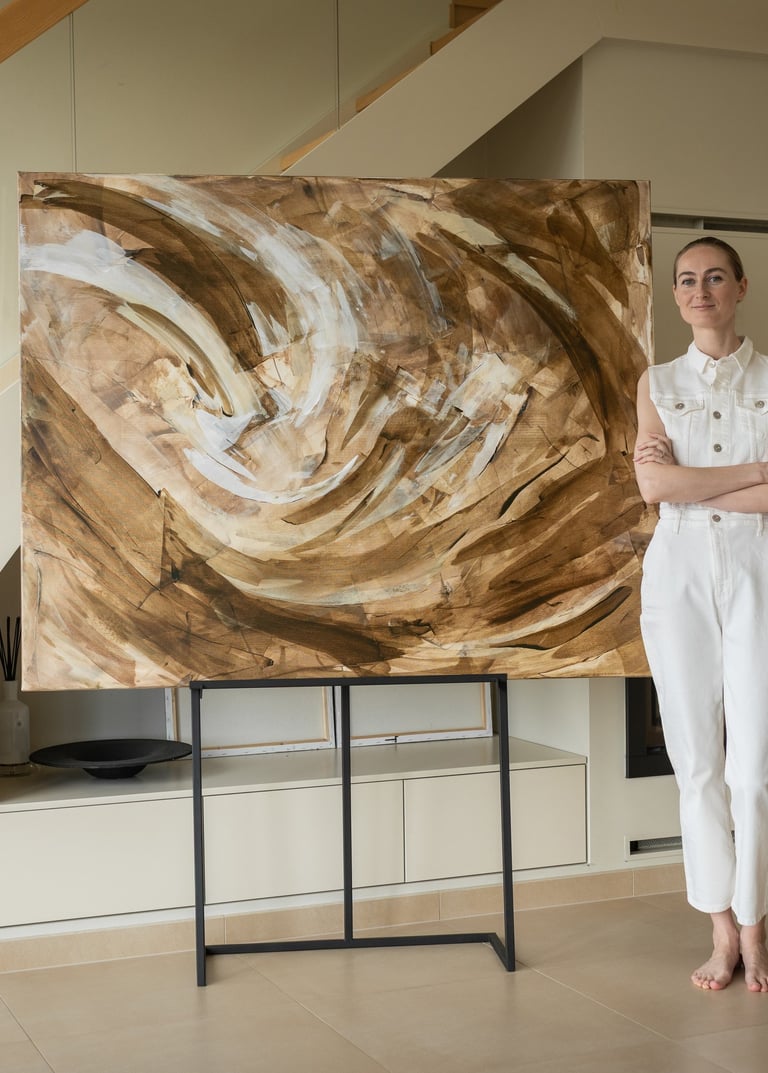 A female artist in a white jumpsuit standing next to her large abstract brown and beige painting on an easel.