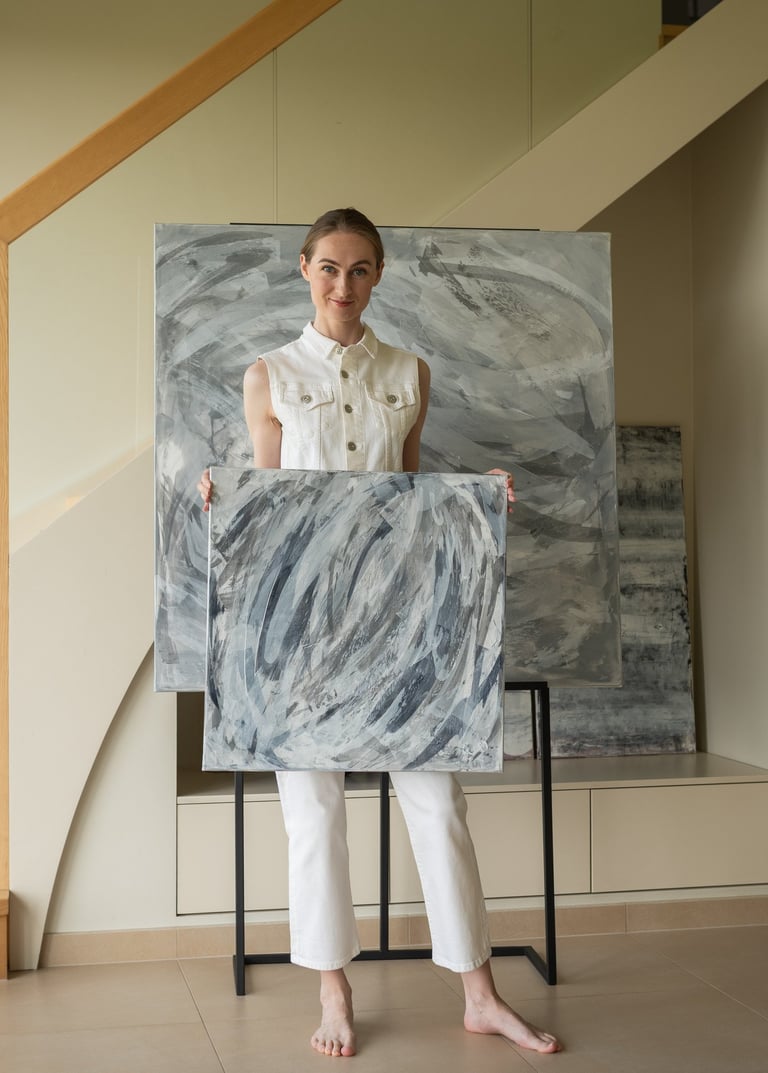 A smiling artist in a modern home holding a grey and white abstract canvas painting near a staircase.