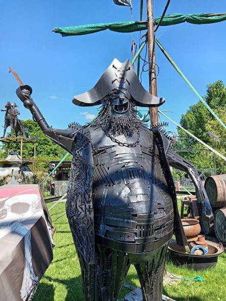 A large black metal pirate sculpture made of scrap metal standing outdoors in front of a ship mast.