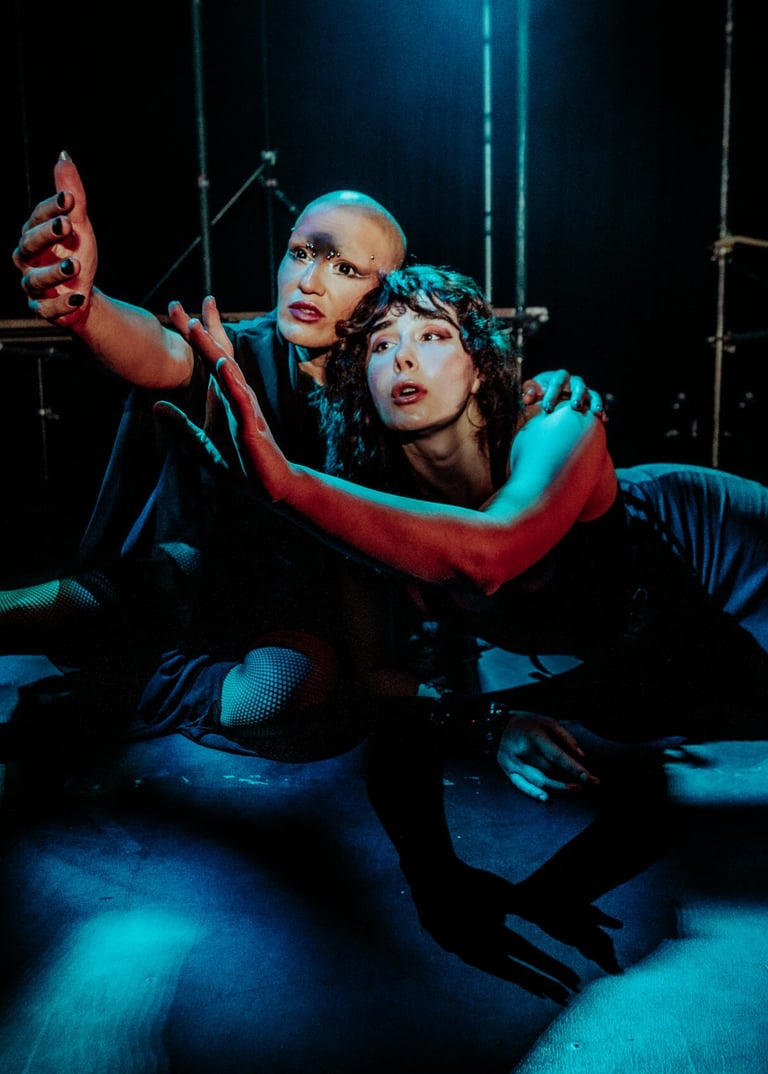 Ila and Coda, bathed in blue light and shadow reach their arms up together, their gazes meeting