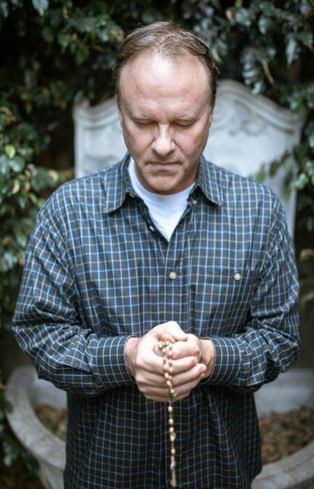 Individual holding a rosary with bowed head in front of a stone monument, surrounded by greenery.