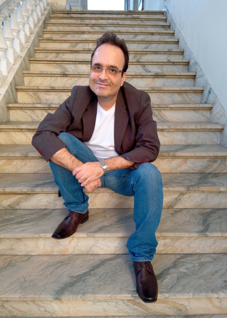 Jornalista Marcelo Dias, sentado à uma escadaria de mármore, de calça jeans, camisa branca e blazer.