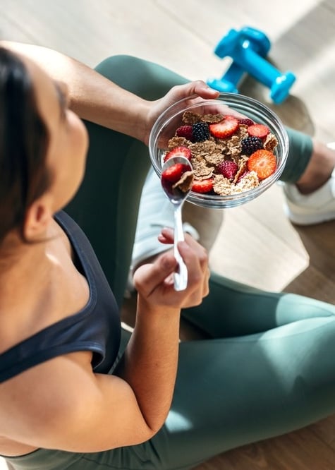 A woman in gym clothes sitting on the floor of a studio with a pair of weights, eating a healthy bowl of fruit and cereal