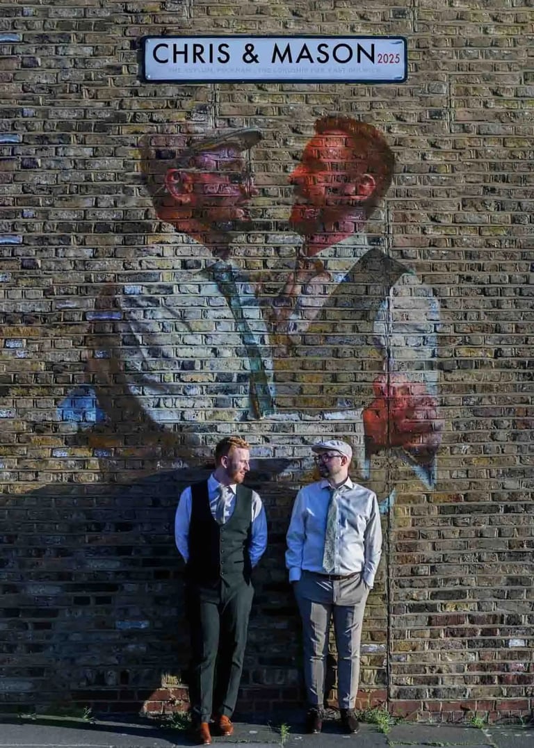 Gay couple posing under a custom street sign and mural of themselves on a London brick wall.