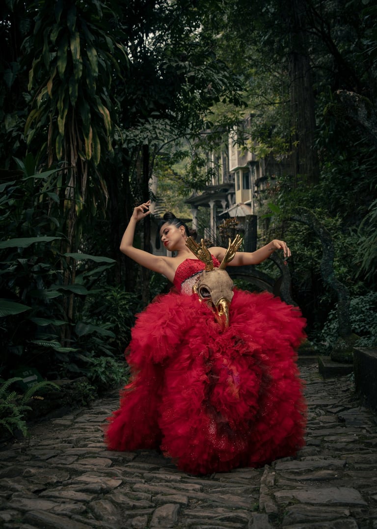 A woman in a dramatic red tulle gown and gold deer skull mask poses in a lush tropical garden.