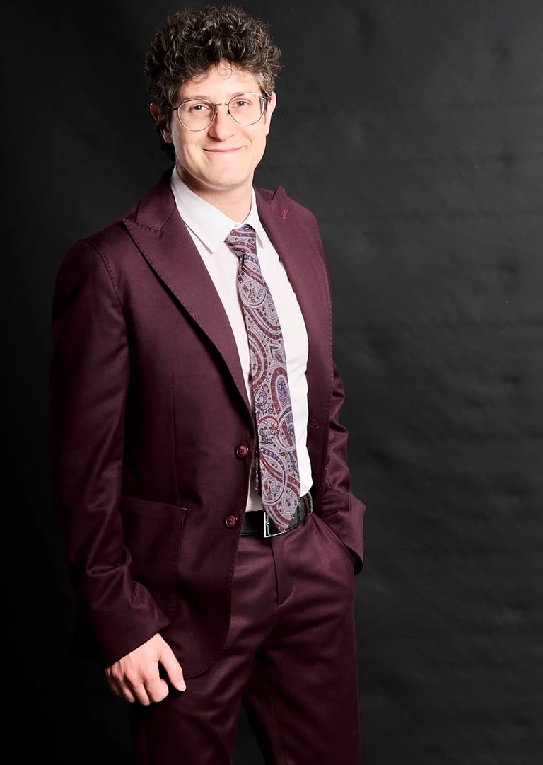Professional portrait of a man in a maroon suit and paisley tie against a black background.