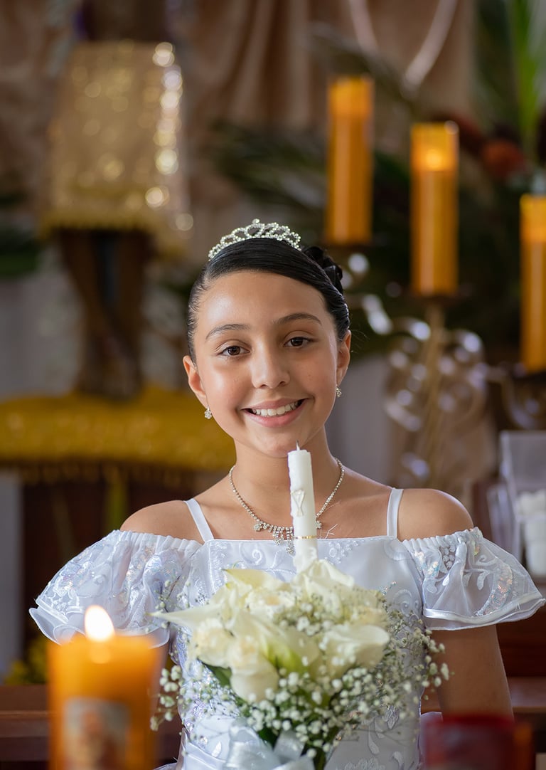 Niña sonriente con vestido blanco de Primera Comunión y tiara sosteniendo una vela ceremonial y un r
