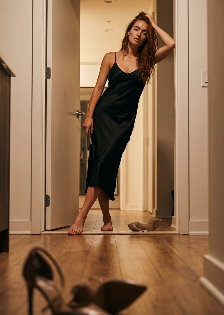 Elegant woman in a black silk slip dress posing barefoot in a doorway with high heel shoes in the foreground.