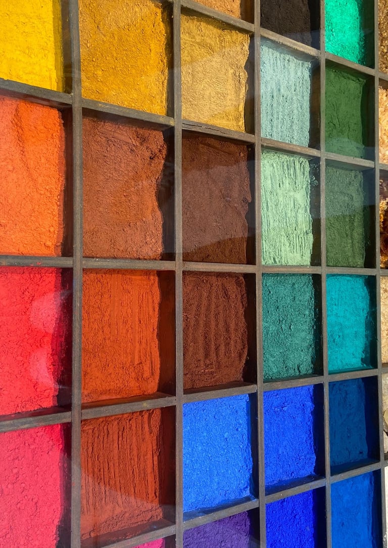 A wooden grid display showcasing a vibrant rainbow of loose powdered pigments and art supplies.