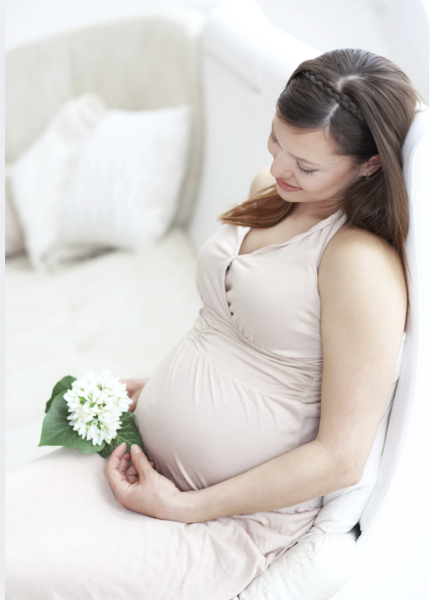 A smiling pregnant woman sitting on a white chair holding white flowers while wearing a beige dress.
