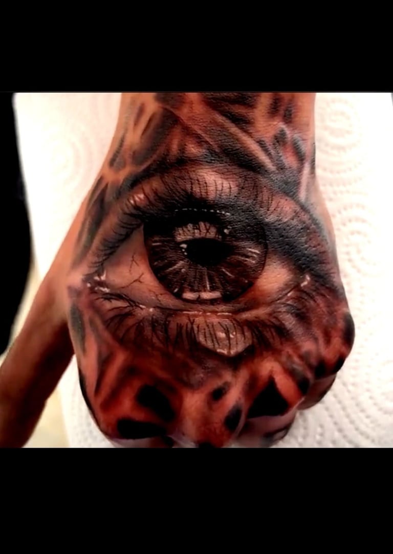 Realistic black and grey eye tattoo on a hand with intricate pupil details and surrounding dark patterns.