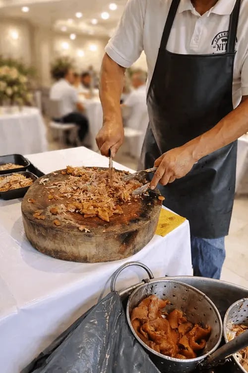 Taquero preparando carnitas en tabla de madera durante un evento, con invitados al fondo.