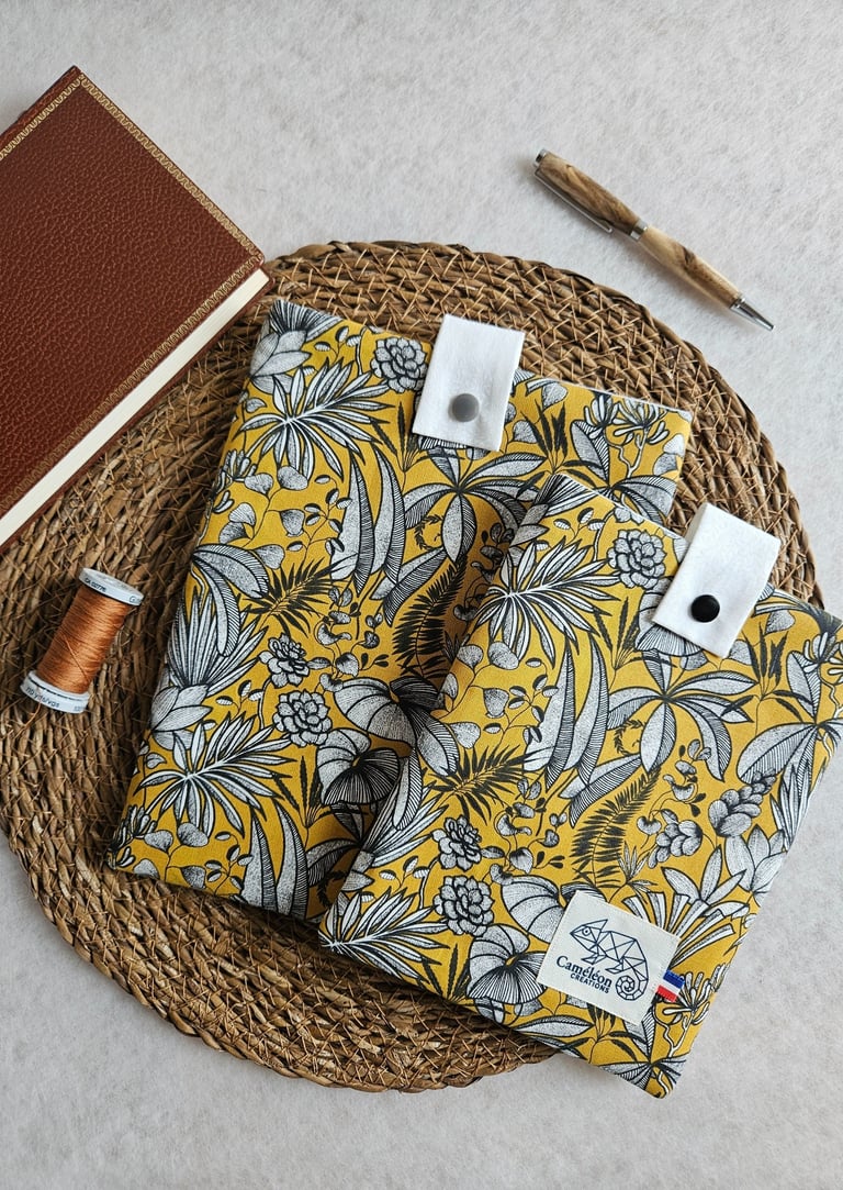 Yellow floral fabric book sleeves with a vintage journal and wooden pen on a woven mat.