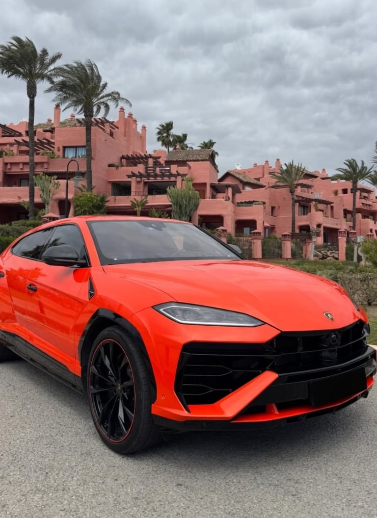 Bright orange Lamborghini Urus luxury SUV parked in front of a pink Mediterranean villa with palm trees.