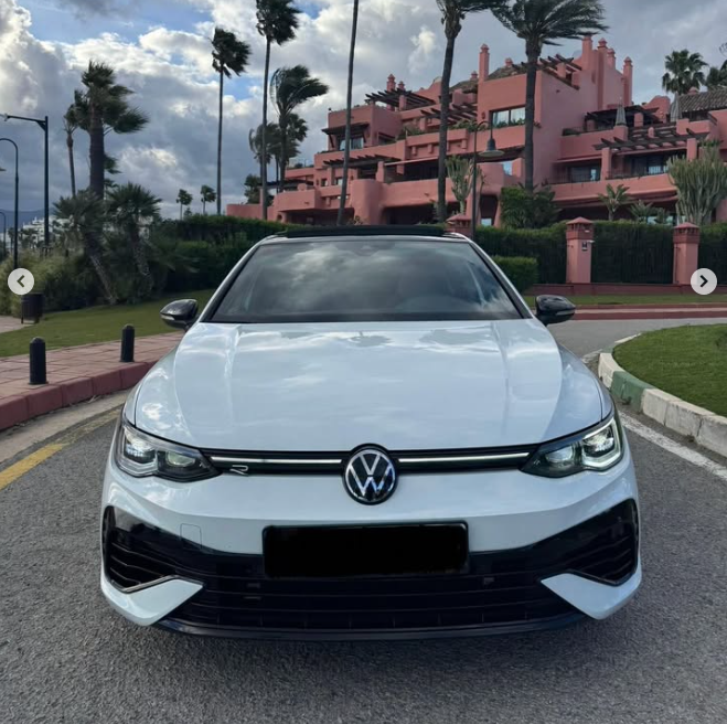 Front view of a white Volkswagen Golf R parked on a coastal road near palm trees and pink buildings.