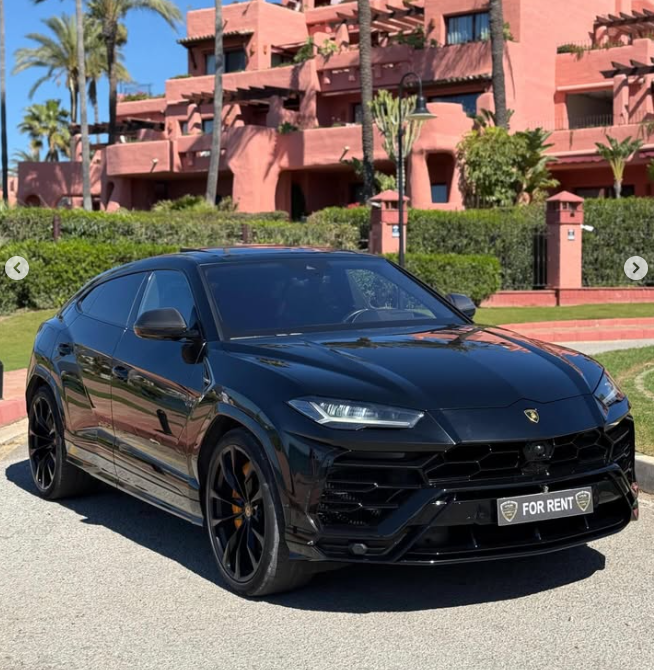 Black Lamborghini Urus luxury SUV for rent parked in front of a Mediterranean resort with palm trees.