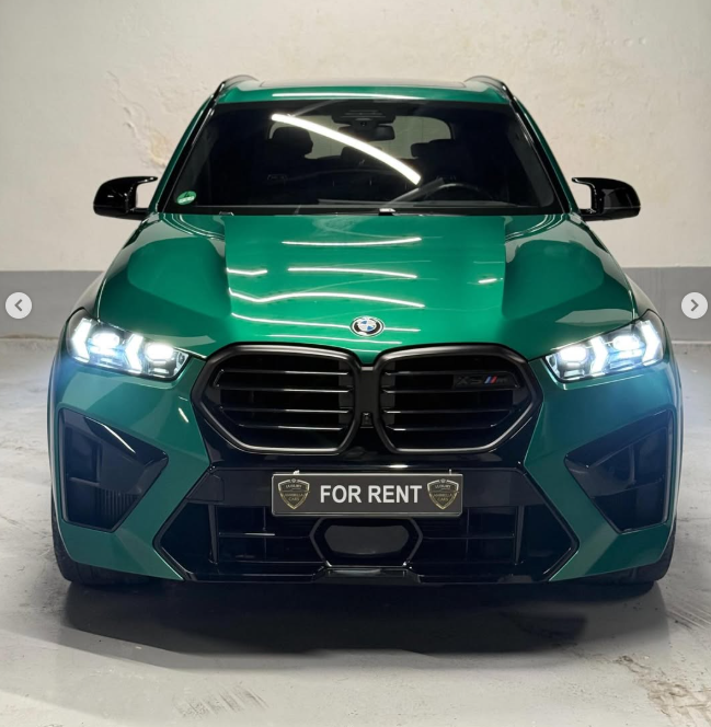 Front view of a luxury green BMW X5 M competition rental car with headlights on in a showroom.