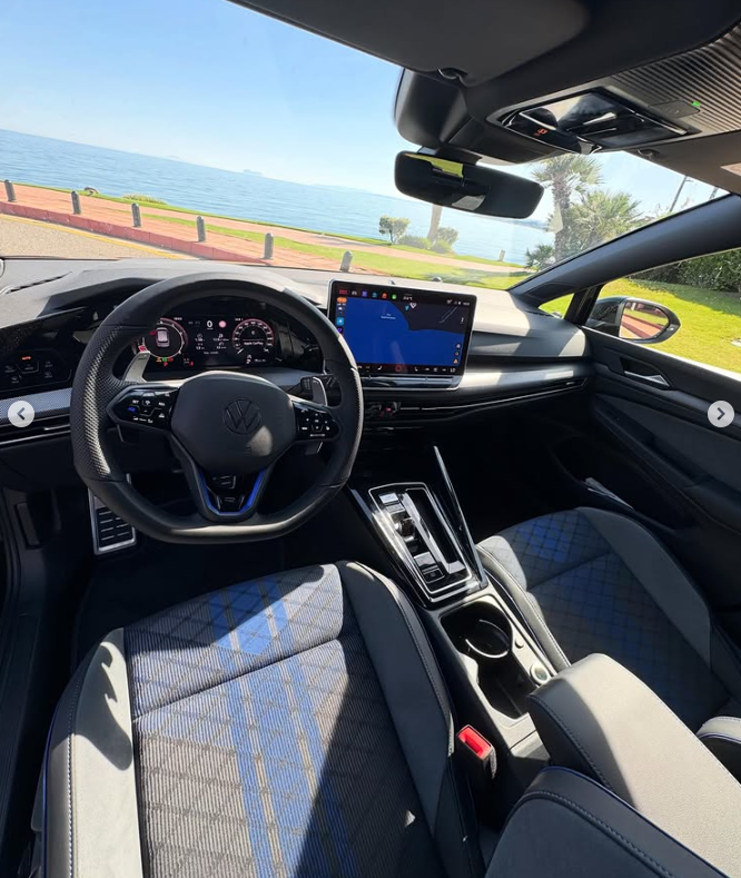 Interior view of a Volkswagen Golf R featuring a digital cockpit and blue plaid seats parked by the ocean.