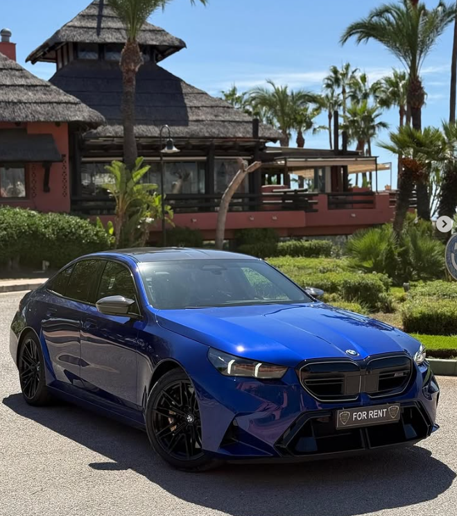 A blue luxury BMW sedan for rent parked in front of a tropical resort with palm trees.