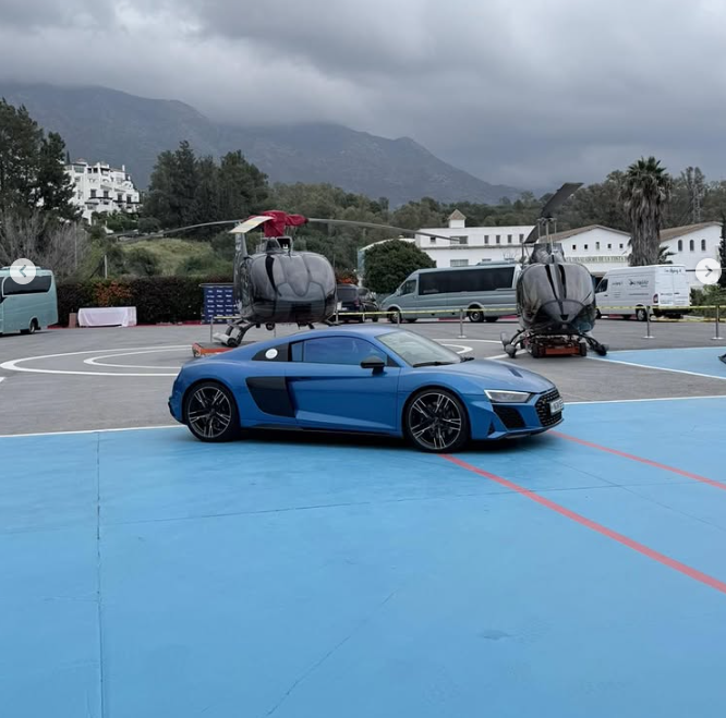 Blue Audi R8 luxury sports car parked on a helipad with black helicopters and mountain scenery.